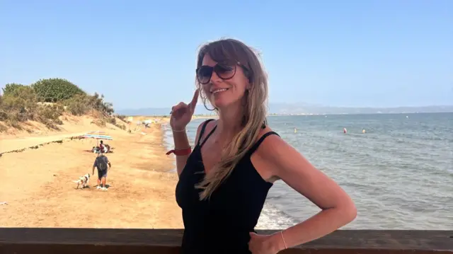 A woman poses with her finger to the sky in front of the sea and a sandy beach as a man walks his dog and people sit under an umbrella.