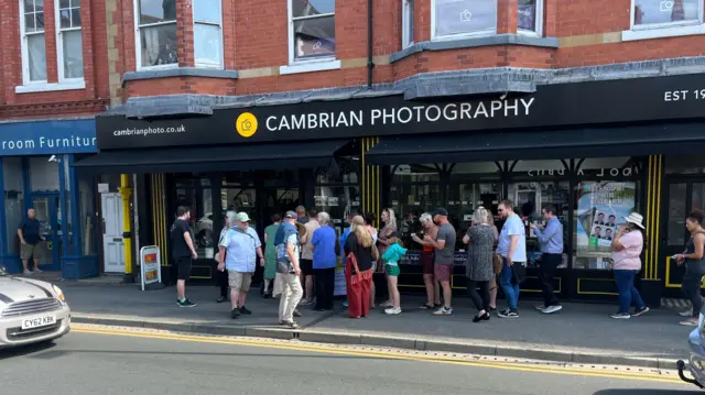 People lining up on a sidewalk outside a photography shop next to a road.