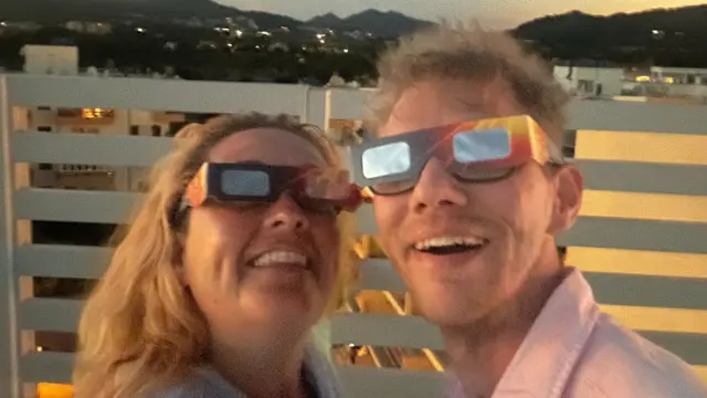 A woman and man stare at the camera wearing eclipse glasses, both are grinning.
