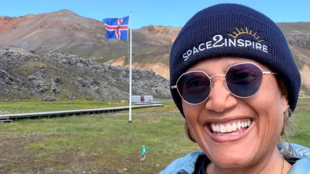 Dr Sian Proctor smiles in a selfie, wearing sunglasses and a hat that reads "space2inspire". An Icelandic flag flies behind her.