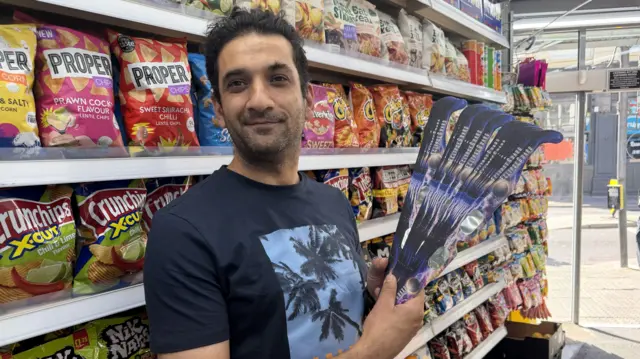 A man smiles at the camera while holding a bunch of cardboard glasses in front of row of crisp packets in a store.