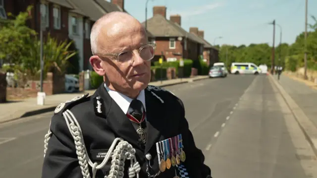 Mark Rowley stands in uniform on a residential street, with his back to the road.