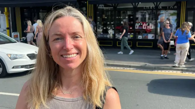 A woman with blonde hair smiles at the camera in front of a road and shopfront with people walking by.