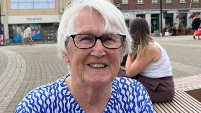 A woman with white hair and glasses wearing a blue and white shirt smiles into the camera