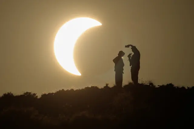 People take photo silhouetted against sky with crescent shaped eclipse visible