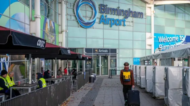A person wearing a dark red coat, yellow rucksack and dragging a wheeled suitcase, walks towards the arrivals entrance at Birmingham Airport.