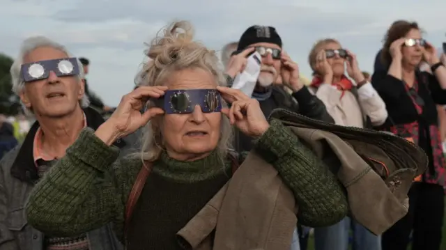 Several people stare at the sky with eclipse glasses on. A woman in the foreground is wearing a green jumper