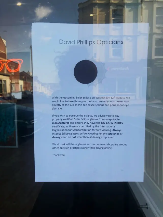 A glass door with a sign on it headlined "David Phillips Opticians" below is a sun and moon diagram showing a partial eclipse and a note warning people not to look directly and the sun and purchase certified solar eclipse glasses