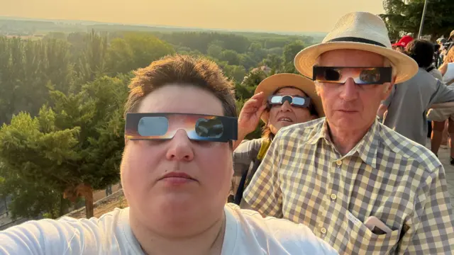 Several people look at the camera while wearing eclipse glasses. The background is sunny and there is a green forest visible.