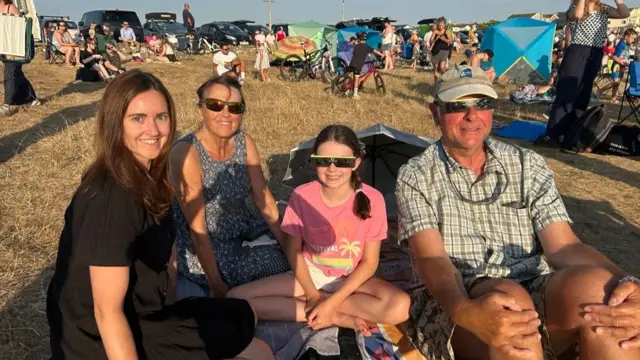 Four people smile while sitting on the ground. The woman on the left is not wearing any glasses, while the mother beside her is wearing normal sunglasses. The child and man are wearing eclipse glasses.