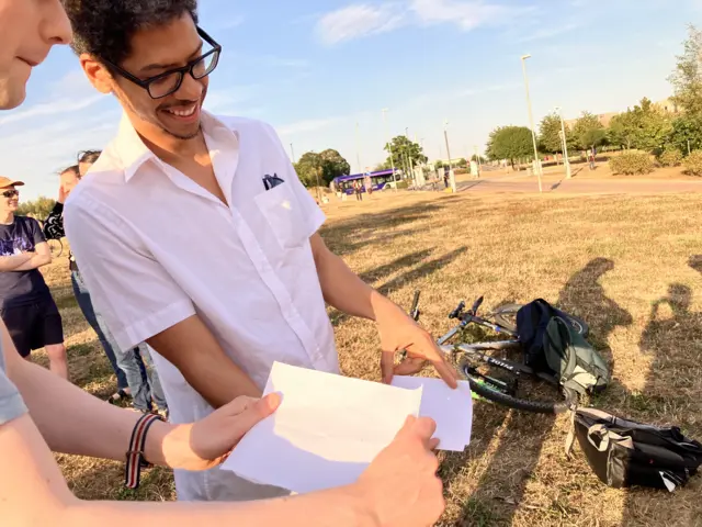 two people hold pieces of paper apart from each other. The top sheet has three pinholes stabbed in it. A tiny projected Sun is visible on the bottom sheet.