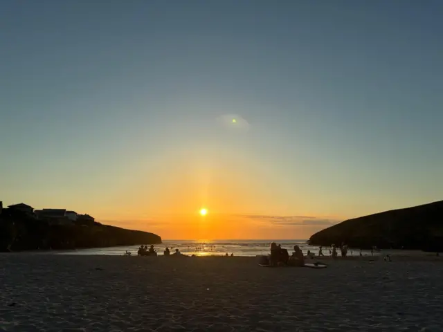 A sunset over a beach between two cliffs jutting out on both sides. People in shadow can be seen sitting on the beach