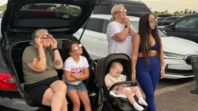 Five people, including an asleep baby, stand and sit at the back of a parked car. The four awake people are looking at the sky wearing eclipse glasses.
