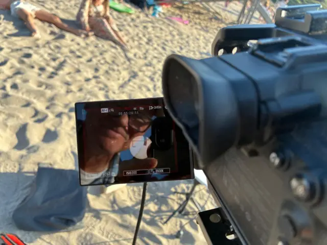 A man looks into the display screen of a camera. He is wearing glasses a white t shift and a blue checked shirt. In the background people are lying on the sand on a beach