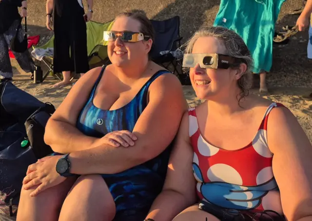 Two ladies sat on the beach wearing solar glasses