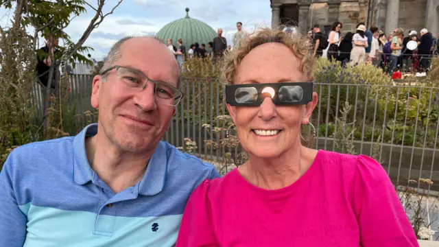 A man and women smile at the camera, the man wearing regular glasses and the woman wearing eclipse glasses