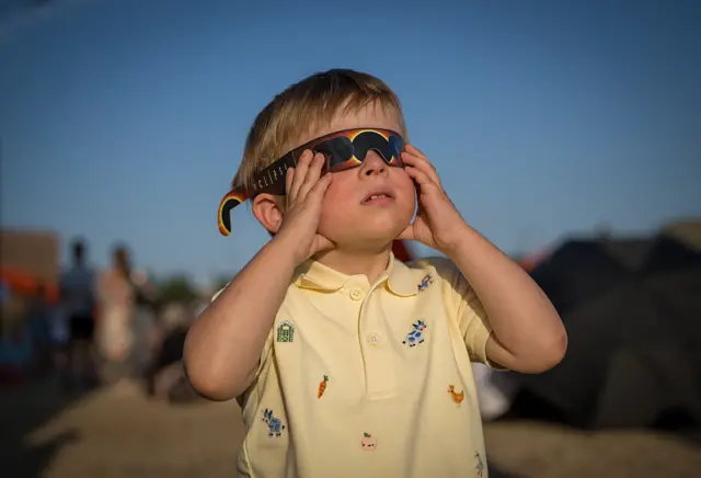 A young boy in a pale yellow polo shirt wears eclipse glasses while looking at the sky