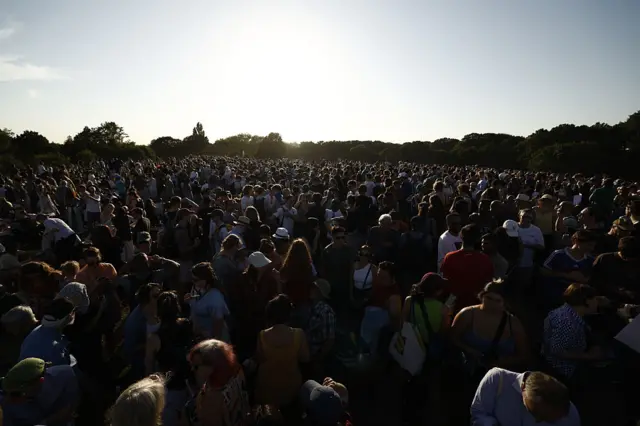 A crowd of people sit in a field with a bright sky overhead.