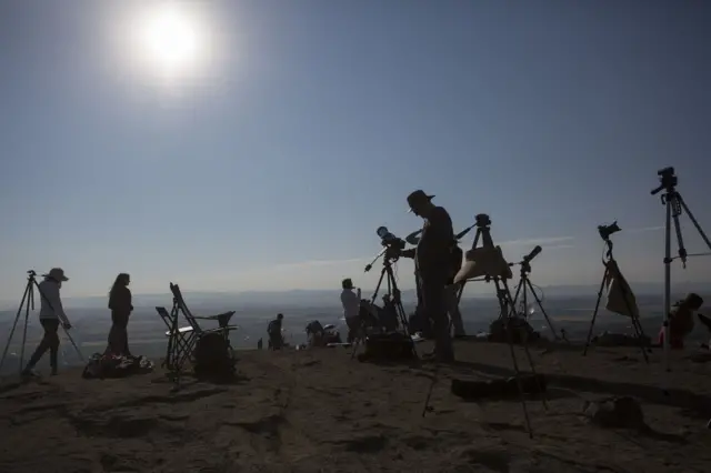 People with telescopes gather on a rocky outcrop high above the background. They are in shadow as the sun is towards the camera