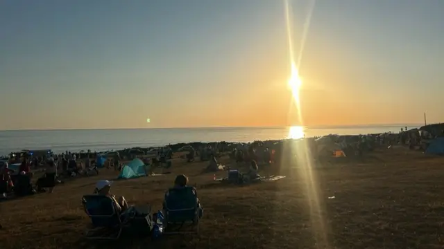 Crowds of people sit on a coast in front of the sea and a bright sky, with the sun in the middle right of the image.
