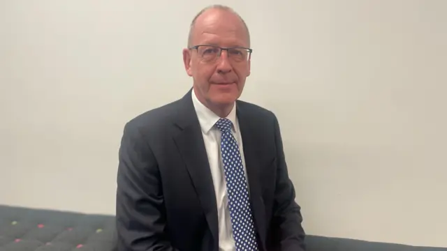 Nick Barton, wearing glasses, a tie, white shirt and black suit jacket, looks at the camera as he sits on a dark grey sofa in front of a white wall.