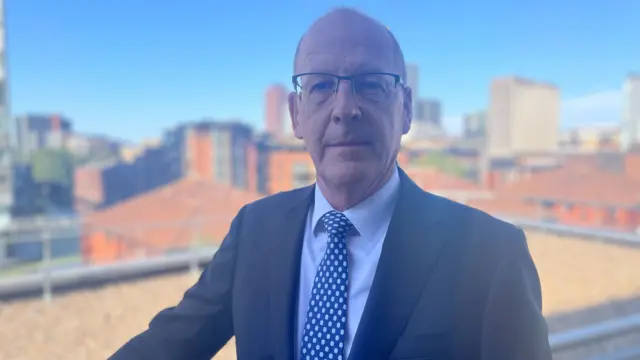 Nick Barton - a bald man wearing glasses, a tie and suit - looks at the camera as he leans on a balcony with a cityscape in the background.