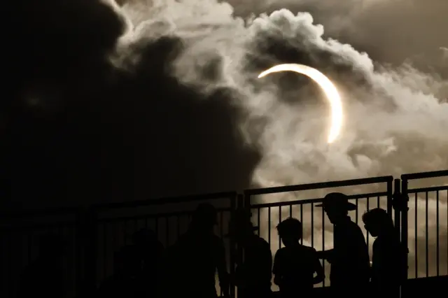 People silhouetted against eclipse with clouds and stand