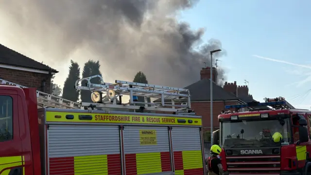 Two fire engines are positioned in front of houses with a large plume of smoke in the backgorund.