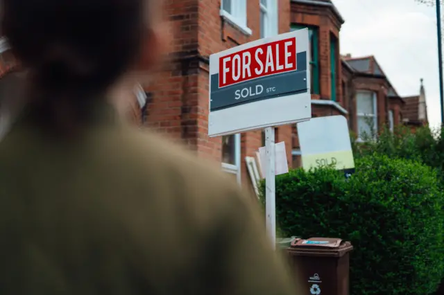 A person stands with their back to the viewer with a for sale sign behind, and a row of redbrick houses behind that.