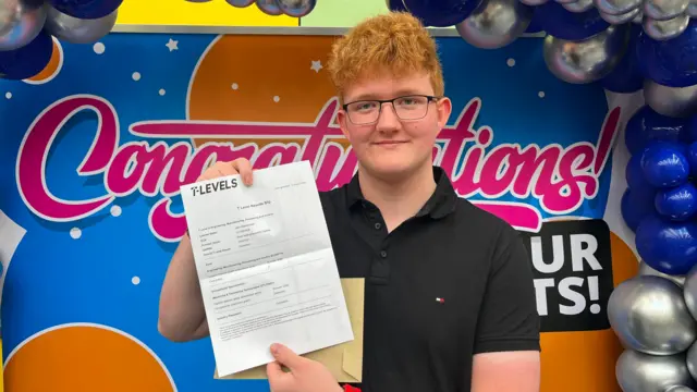 Jake Myerscough wears a black polo shirt and holds up a T Levels results paper