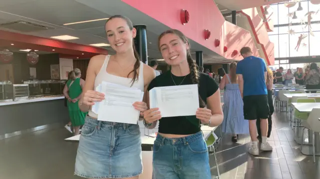 Two girls opening their A level results smiling, one with a white top and blue denim skirt, the other with a black top and blue denim shorts