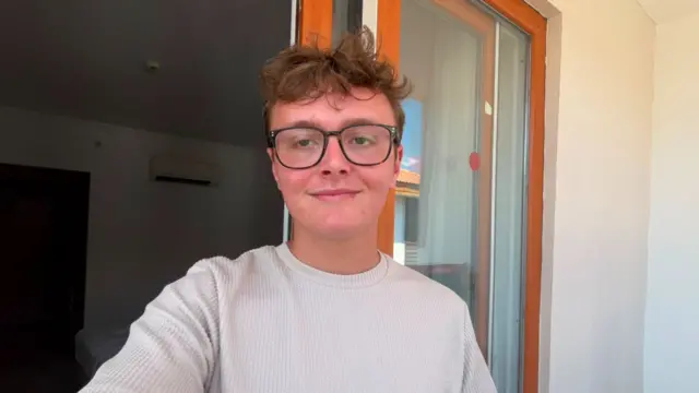 Selfie of a young man with short, light brown hair and black glasses taken in front of a glass-panelled door