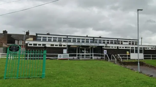 A single-storey building with a patch of lawn in front. There is a green gate positioned at the bottom of a sloped section of the lawn. There are steps leading up to the entrance of the building.