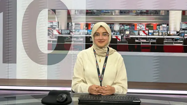 Journalist sitting behind a desk with the BBC newsroom in the background.