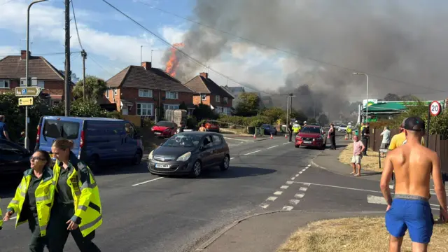 Flames are seen erupting from a house of the left of a busy street. The house is situated opposite a BP petrol station and police have shut the bottom half of the road.