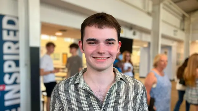 Stephen is seen holding his results paper in a large indoor hall, standing in front of a blurred background with several people. A vertical sign reading “BE INSPIRED” is visible on the left. The venue has high ceilings and bright lighting.