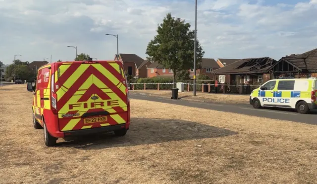 A yellow and red van is parked on a dry patch of grass. A police van is parked in the background in front of the damaged bungalows.