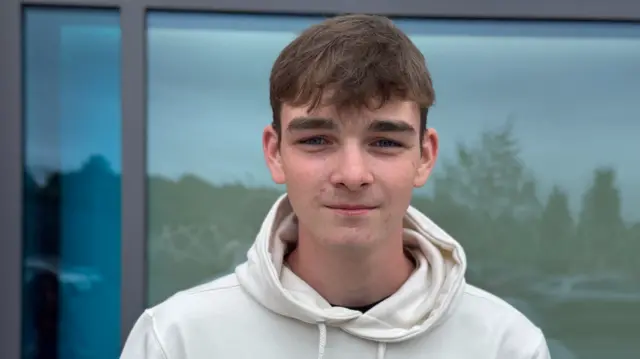 Jack Brannigan smiles as he holds up his results. He is wearing a white hoodie