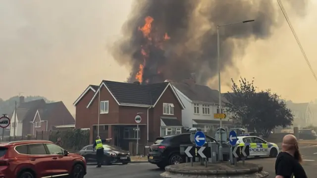 Flames tower over a row of two storey houses while police officers direct people away from the scene on a small roundabout.