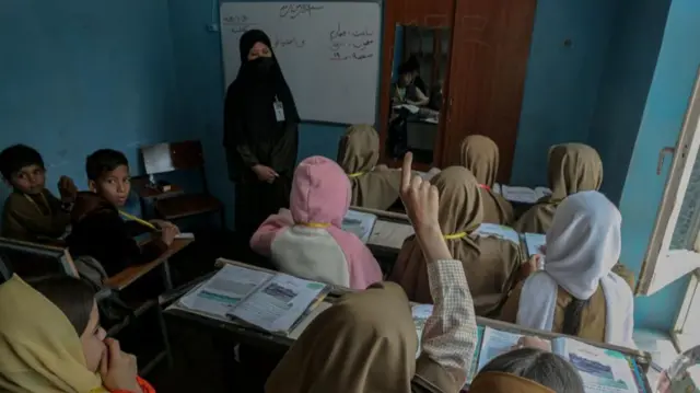 A group of young students are pictured from behind in a classroom, with boys segregated to the left and girls on the right. One of the girls has her hand raised.