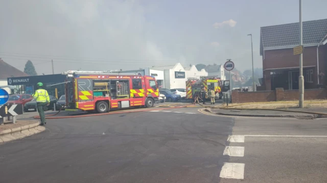Multiple fire engines are parked along a street with a Renault car dealership on one side and red brick houses on the other. Hose pipes are laid out across the road and fire fighters are dotted around the area. Smoke fills the sky and appears to be coming away from behind the car dealership.