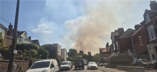A large plume of smoke can be seen in the distance, billowing above a residential street in the foreground with cars parked either side.