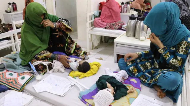 Two women in full veil coverings are shown on a bed with babies lying in front of them. There are various papers on the bed.