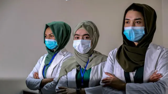 Three women in white medical coats, face masks and headscarves are shown standing with their arms crossed.