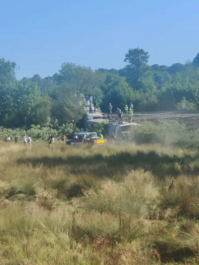 A train on its side. People are stood on top of the derailed carriage.