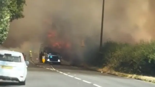 A light coloured vehicle is parked at the side of a road while a fire engine engulfed in smoke and flames is seen in the distance with firefighters nearby.