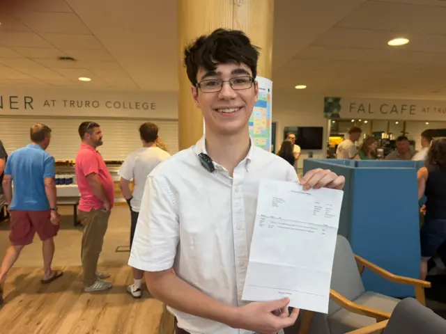 Nick Basher smiles as he holds up his results as he speaks to the BBC. He is wearing a white shirt