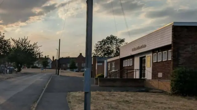 A single-storey brick building with white wooden cladding across the top is at the side of a road.) The photograph appears to have been taken at sunset.