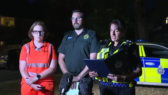 A Network Rail representative in orange luminous clothing stands beside an ambulance service representative in a dark green uniform, with a police officer beside him reading from a folder.