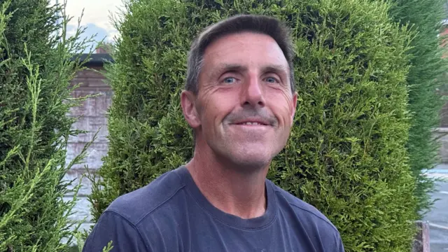 Robert Windsor looks into the camera, he has short, dark hair and slightly smiles, showing his teeth, in front of a green bush